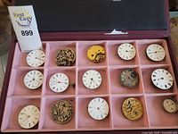 Photo showing 15 different watch and clock dials and movements arranged in a pink divided tray.