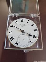 Front view of the dial in plastic case showing Roman numeral enamel dial and hands