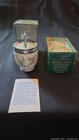 Photo of one small egg coddler standing upright with metal lid and floral decorated ceramic body next to its original green box and instruction leaflet on a black background.