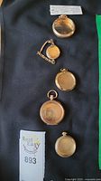 Five gold-colored pocket watch and wrist watch cases laid out on black cloth, with a note and lot number 893 visible.