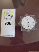 Howard & Co watch movement placed next to auction lot number card, showing the face of the watch dial with Roman numerals.