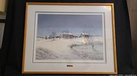 Framed print showing a winter rural scene with snow-covered houses and a pond under a dark blue sky.