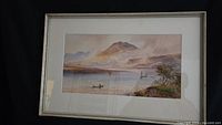Framed watercolor painting showing a lake with mountains and boats, signed by artist.