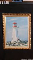 Frontal view of framed painting showing lighthouse and surrounding rocks with three people at lighthouse base.