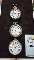 Three pocket watches displayed vertically on a dark background showing front dials and cases.