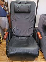 Front view of black leather massage recliner showing padded seat, backrest, and wooden armrests with some scuffs.