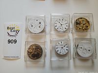 Six pocket watch movements and dials in clear plastic boxes arranged in two rows
