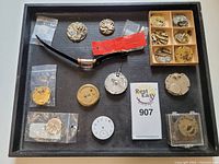 Tray containing various watch parts including multiple mechanical movements, watch dials, a wristwatch with black strap, and small components in plastic bags.