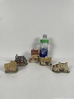 Four small David Winter collectable cottages arranged in front of a water bottle for scale, showing detailed textures, roof styles, and miniature windows.