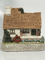 Front view of the first collectible cottage featuring a brown shingled roof, white chimney, stone base, and detailed foliage.