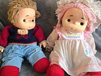 Photo showing both dolls seated on a couch holding hands, showing boy doll's red sweater and blue pants and girl doll's white dress.
