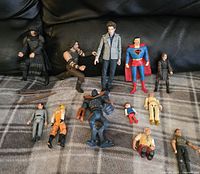 Overview of assorted toy figures including Superman, a man in gray jacket, and other action figures and dolls on a plaid surface, against a black background.