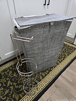 Gray woven laundry basket standing with attached hinged lid closed; silver metal toilet paper holder next to it.
