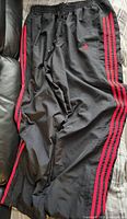 Photo shows front view of black Adidas men's windbreaker athletic pants, size XL, with three red stripes running down the outer thigh of each leg and drawstring waistband.
