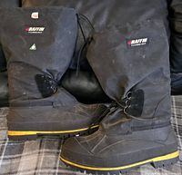 Side view of paired black BAFFIN boots with yellow sole stripe and drawstring closure at calf, showing brand patch.