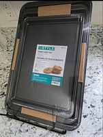 Three brand new black nonstick cookie sheets stacked and factory wrapped in clear plastic with cardboard spacers.