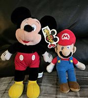 Photo showing brand new Mickey Mouse plush toy and Mario plush toy side by side on a dark couch.