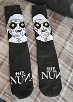 Front view of both black tube socks showing the graphic of the Nun character with yellow eyes and a necklace cross, and the text 'THE NUN' on the foot area.