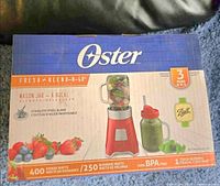 Front view of Oster Fresh Blend-n-Go Mason Jar Blender box showing blender with mason jar filled with fruit and red lid with straw.