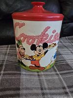 Front view of round white ceramic cookie jar with colorful Disney characters Mickey Mouse, Minnie Mouse, Goofy, holding plates of cookies, with "Cookies" text and red lid.