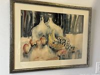 Framed abstract watercolour still life painting with stylized bottles, fruit, and bowl on table.