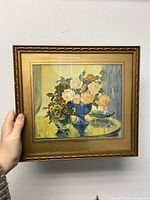 Gilt framed floral art print with yellow and white roses in a blue vase on a reflective round surface, held up for viewing
