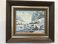 Framed winter landscape oil painting showing composition with snow, trees, rocks, and water under a partly cloudy sky.