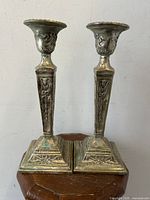 Front view of two silver plated Gothic Victorian candlesticks showing raised ram head and flag detail on tapered stems and square bases