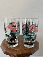 Front view of 2 glasses showing colorful enameled reindeer and mountain landscape design with pine trees.