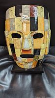 Front view of the life size Aztec death mask sculpture showing the multicolored polished stone mosaic and eye and mouth cutouts mounted on a black display base.