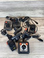 Photo showing a lot of seven Atari controllers, including five joystick controllers mostly black with orange buttons, one joystick missing top casing, and two paddle controllers with orange buttons and silver top faceplate. All have cables attached.