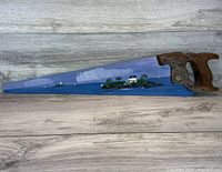 Entire view of a hand saw with a detailed painted scene of a Maine lighthouse island and boat under a cloudy sky on the saw blade, wooden handle visible.