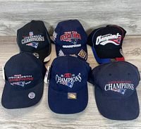 Six New England Patriots championship hats in navy blue and black, displayed on a wooden surface. Some hats have tags attached indicating new condition.
