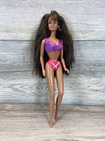 Front view of vintage Barbie doll with brunette hair, purple crop top and pink bikini bottoms on wooden floor background.