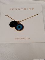 Jenny Bird necklace displayed on branded card showing two gold tone pendant charms including a blue evil eye design and a round charm with "JENNY BIRD" engraving.