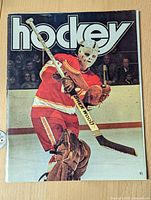 Front cover of 1973 hockey magazine showing goalie in red and white uniform and protective mask holding Sher-Wood hockey stick during game action.