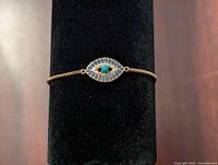 Front view of the gold tone evil eye bracelet displayed on a black stand, showing the blue and turquoise stones on the charm.