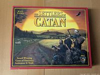 Front of the Settlers of Catan game box showing the game's title and artwork depicting settlers on a rural path with a sunset background.