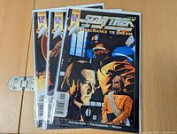 Three Star Trek The Next Generation comic books in plastic sleeves with cardboard backs arranged side by side on a wooden surface.