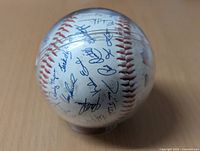 Photo showing part of the baseball with multiple blue ink signatures of Toronto Blue Jays players, white with red stitching inside a clear protective case.
