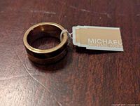 Michael Kors gold-tone band ring with tag showing brand logo, placed on a wooden surface.