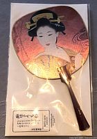 Full view of Japanese hand fan with Geisha image, iridescent finish, dark red handle, in clear plastic packaging