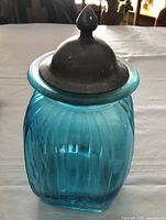 Front view showing turquoise blue fluted square glass jar with metal dome lid topped with acorn-shaped handle.