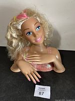 Barbie styling head with blonde curly hair, blue eyes, and pink lips. Upper torso and hands visible with sparkly pink top.