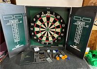 Full view of open wooden cabinet containing Narwhal Kingsbridge dart board with chalk score boards on both doors, multiple darts and accessories laid out on table.