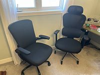 Two black office chairs placed side by side in front of a window, one with a high mesh back and headrest, the other with a cushioned backrest