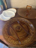 Photo showing the large amber glass plate with ornate pressed floral and geometric design, white three-compartment dish with gold trim, and two amber glass utensils.