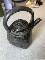 Front and side view of the dark grey pottery teapot showing the textured surface, angular body, and tiered lid with a curved handle.
