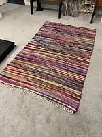 Full view of the rainbow rag rug laid out on a carpeted floor showing multicolor horizontal stripes and fringes at one end.