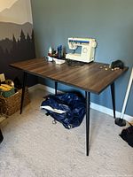 Front angle view of the rectangular dining table showing wood grain top and diagonal black legs, with a sewing machine on top and various small items.